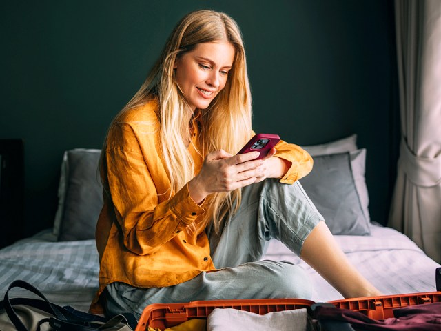 Person sitting on bed looking at phone next to suitcase