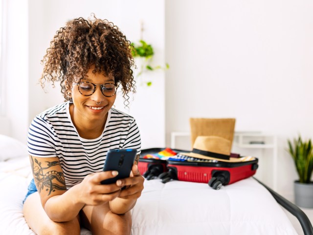 Person sitting on bed looking at phone next to open suitcase