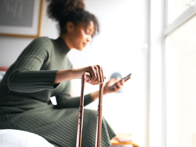 Traveler sitting on hotel bed holding luggage handle and using phone