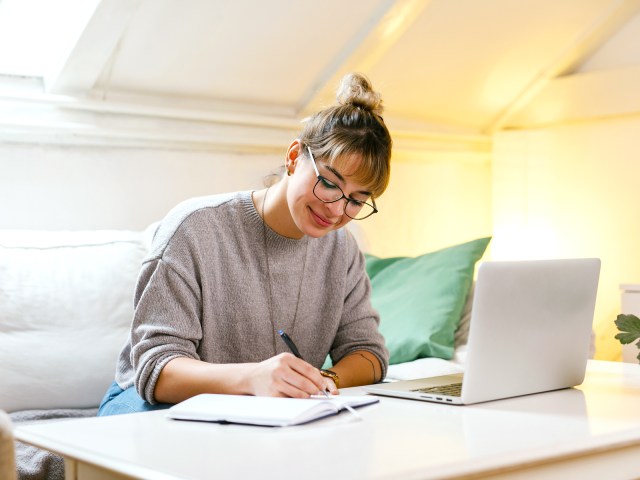 Person sitting at table with laptop computer and writing in notebook