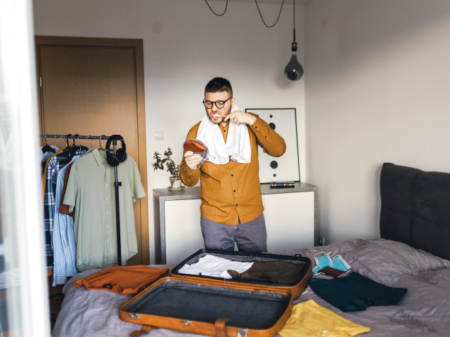 Man brushing teeth and holding mirror while standing next to suitcase on bed