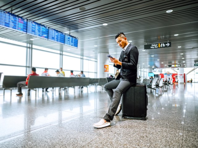 Passenger sitting on suitcase checking phone in airport terminal