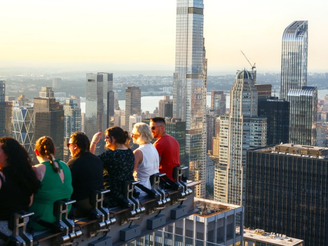 Tourists sitting on the Beam attraction at 30 Rock overlooking NYC skyline